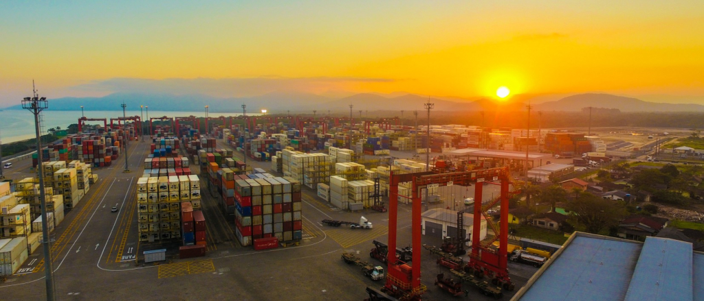 Vista aérea do Porto de Itapoá, em Santa Catarina, ao pôr do sol, com grandes pilhas de contêineres coloridos organizados no pátio, guindastes portuários vermelhos e caminhões circulando entre as áreas de carga. Ao fundo, o mar e montanhas aparecem no horizonte.