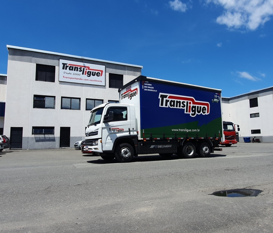 Caminhão elétrico da Transligue estacionado no pátio em frente à sede da empresa. O baú azul e verde destaca "zero emissões", sendo uma opção sustentável para o transporte rodoviário.
